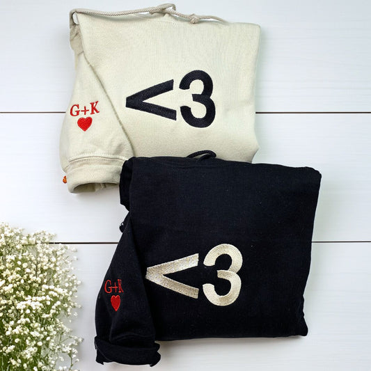 Matching Valentine's Day hoodies in black and cream with embroidered heart and '<3' design, personalized initials on sleeve.