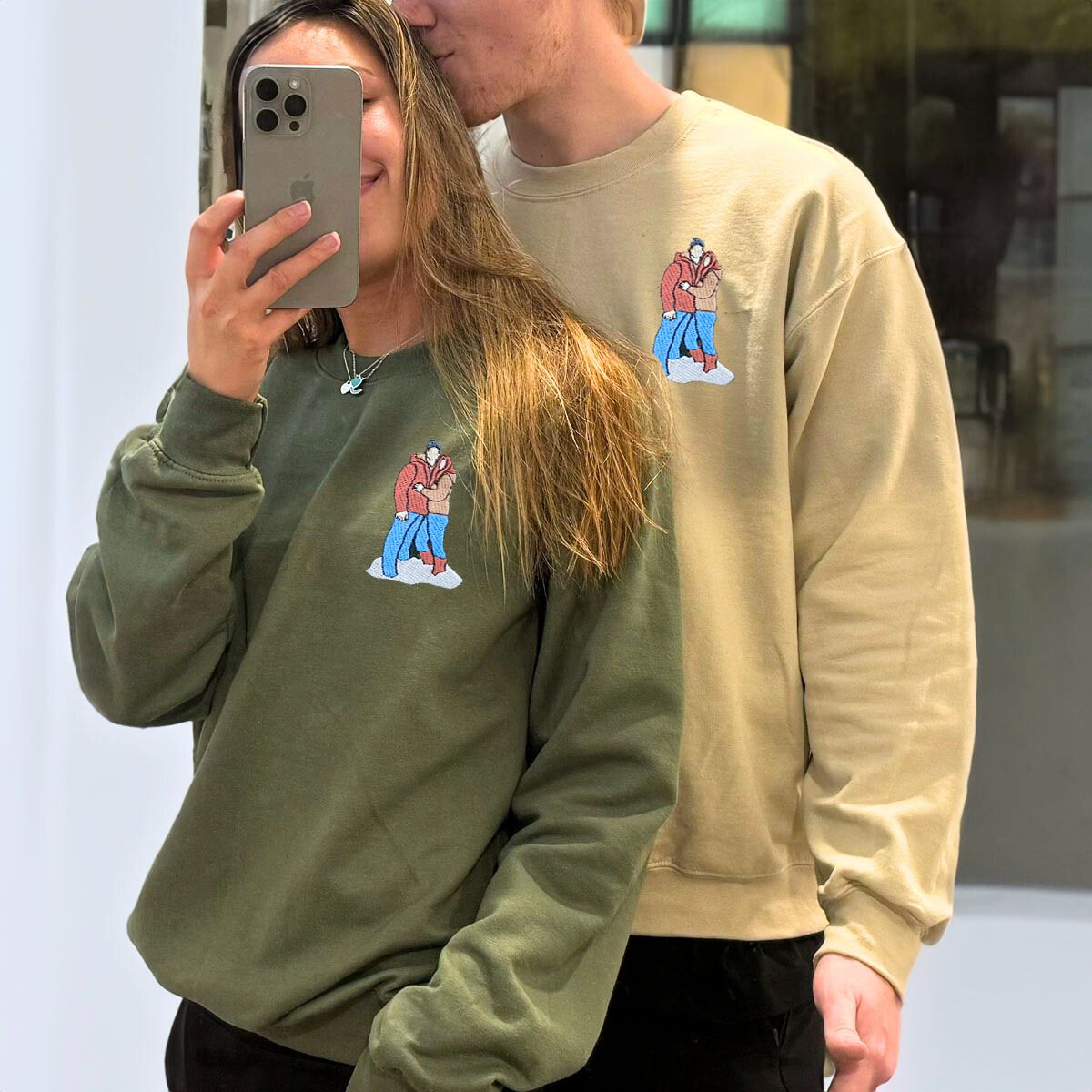 Affectionate couple taking a mirror selfie in beige and green Embroly sweatshirts with custom winter scene embroidery.