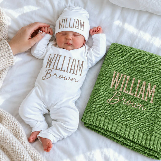 Sleeping newborn in a personalized white bodysuit and hat beside a matching embroidered green knit blanket, a gift idea for a baby shower.