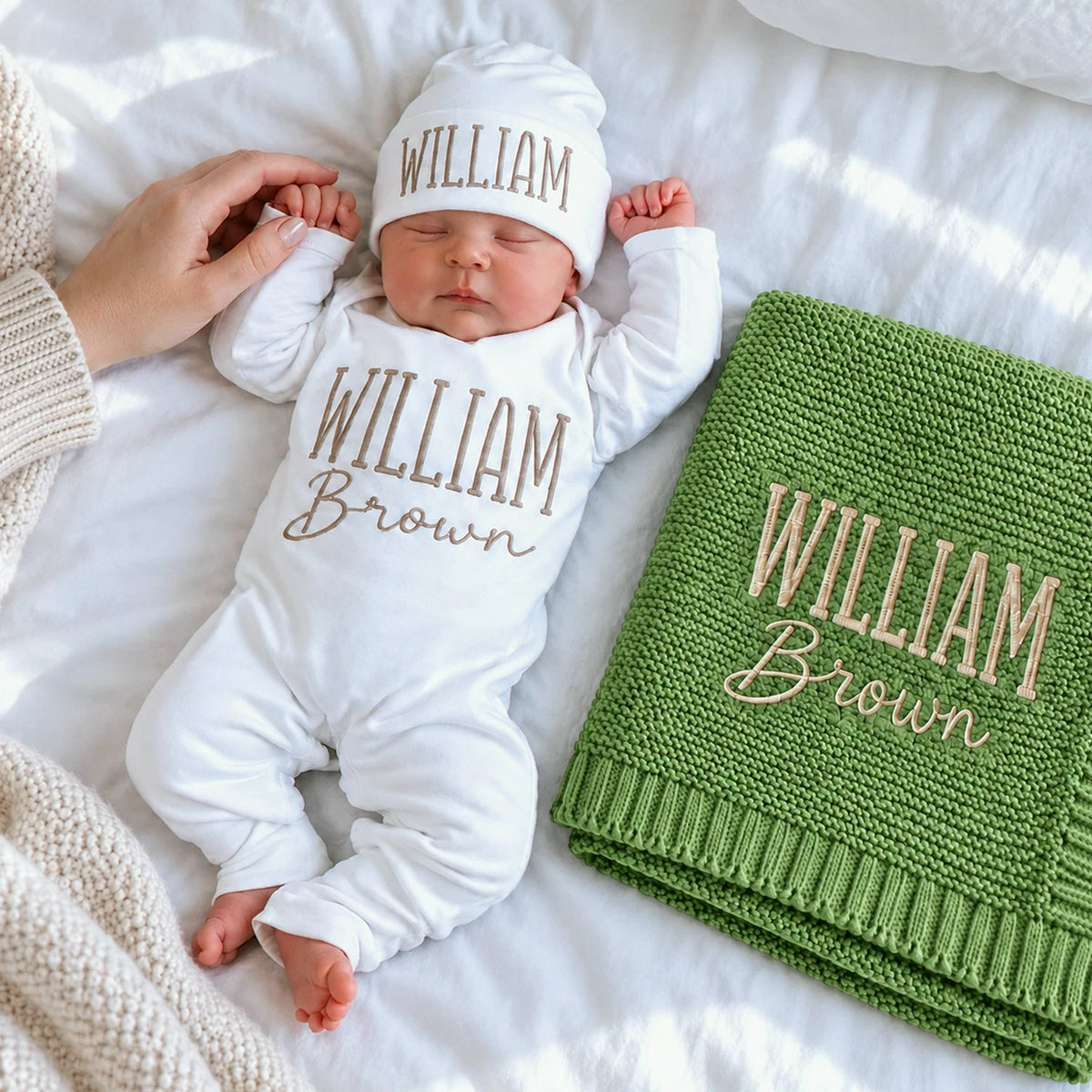 Sleeping newborn in a personalized white bodysuit and hat beside a matching embroidered green knit blanket, a gift idea for a baby shower.