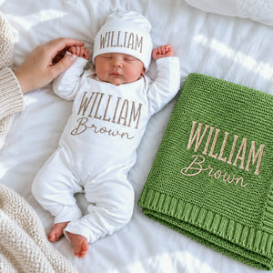 Sleeping newborn in a personalized white bodysuit and hat beside a matching embroidered green knit blanket, a gift idea for a baby shower.