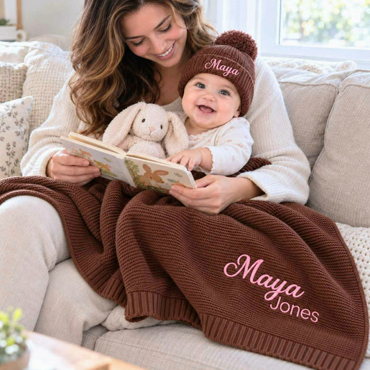 Mother reading with her baby under a chocolate brown knit blanket embroidered with pink names, with a matching beanie, a personalized christening gift.