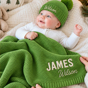 Smiling baby with a personalized green knit blanket embroidered with two names and a matching pom-pom beanie, a gift for baby’s first Christmas.