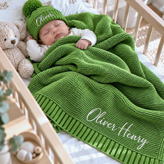 Sleeping baby in a personalized sage green knit blanket and matching embroidered pom-pom beanie, with a parent’s hand nearby, a gift for welcoming a newborn.