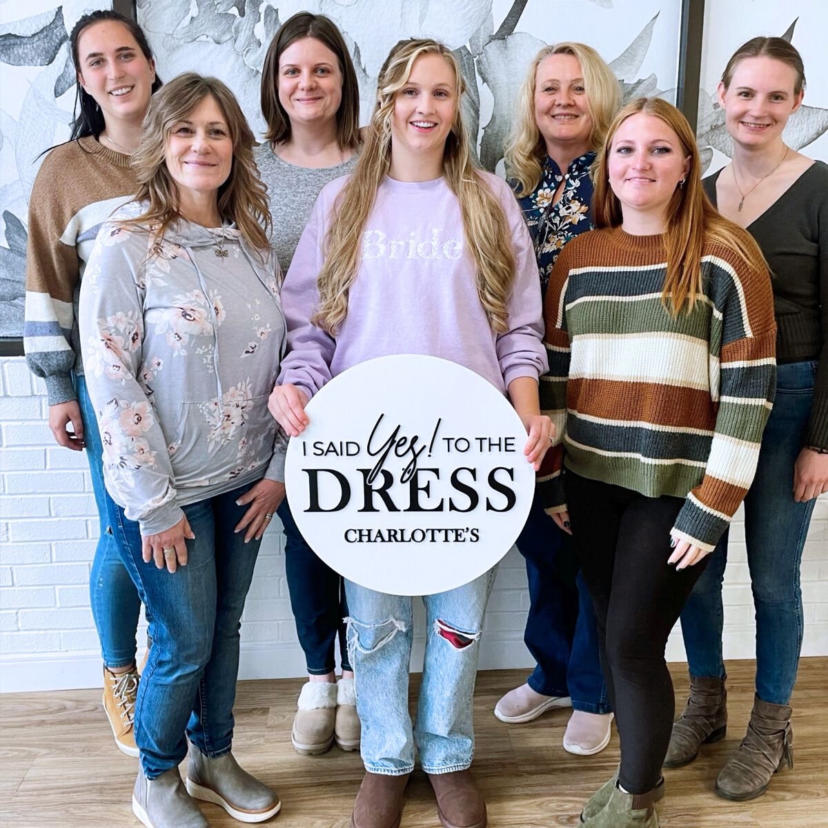 Bride in a pastel sweatshirt with friends, holding an 'I Said Yes to the Dress' sign.