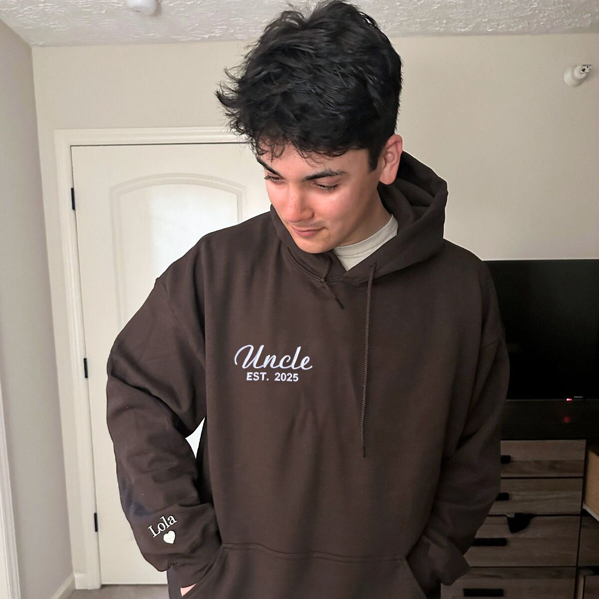 Young man in a brown 'Uncle Est. 2025' hoodie looking down, cozy and warm fabric.