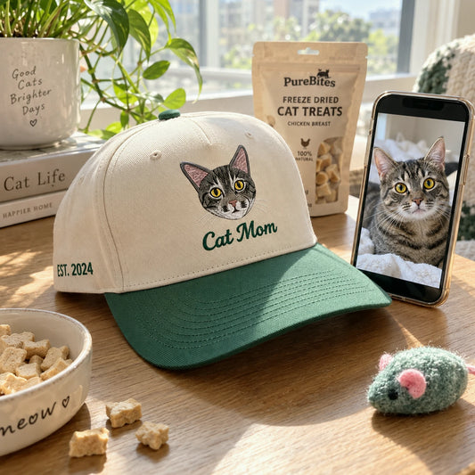 A beige baseball cap with a green brim is displayed on a wooden table, featuring an embroidered cat face and the words 'Cat Mom'. A bowl of cat treats, a pouch of PureBites freeze dried cat treats, and a smartphone showing a cat photo are arranged around it in a bright, sunlit home setting.