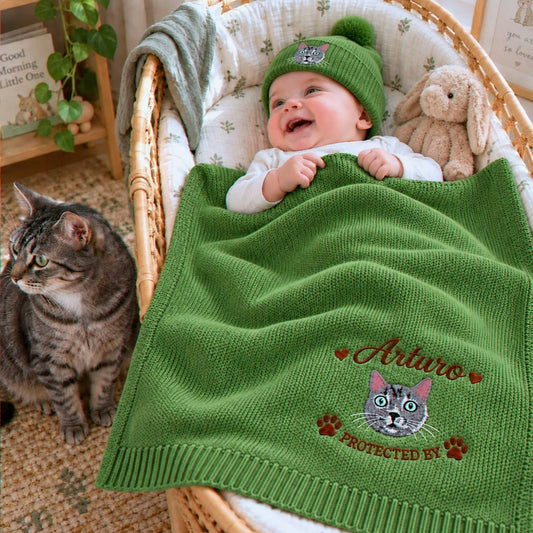 A personalized green knit baby blanket and hat set featuring detailed cat embroidery. The blanket has a custom name and a cat face with "PROTECTED BY" text, ideal as a unique baby shower or new baby gift