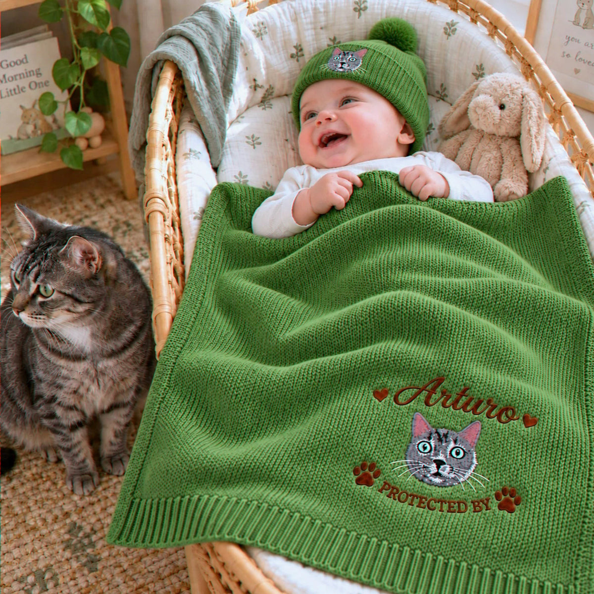 A personalized green knit baby blanket and hat set featuring detailed cat embroidery. The blanket has a custom name and a cat face with "PROTECTED BY" text, ideal as a unique baby shower or new baby gift