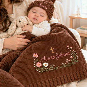 Smiling baby beside a chocolate brown knit blanket embroidered with a floral cross and personalized name, wearing a matching beanie, a baptism gift.