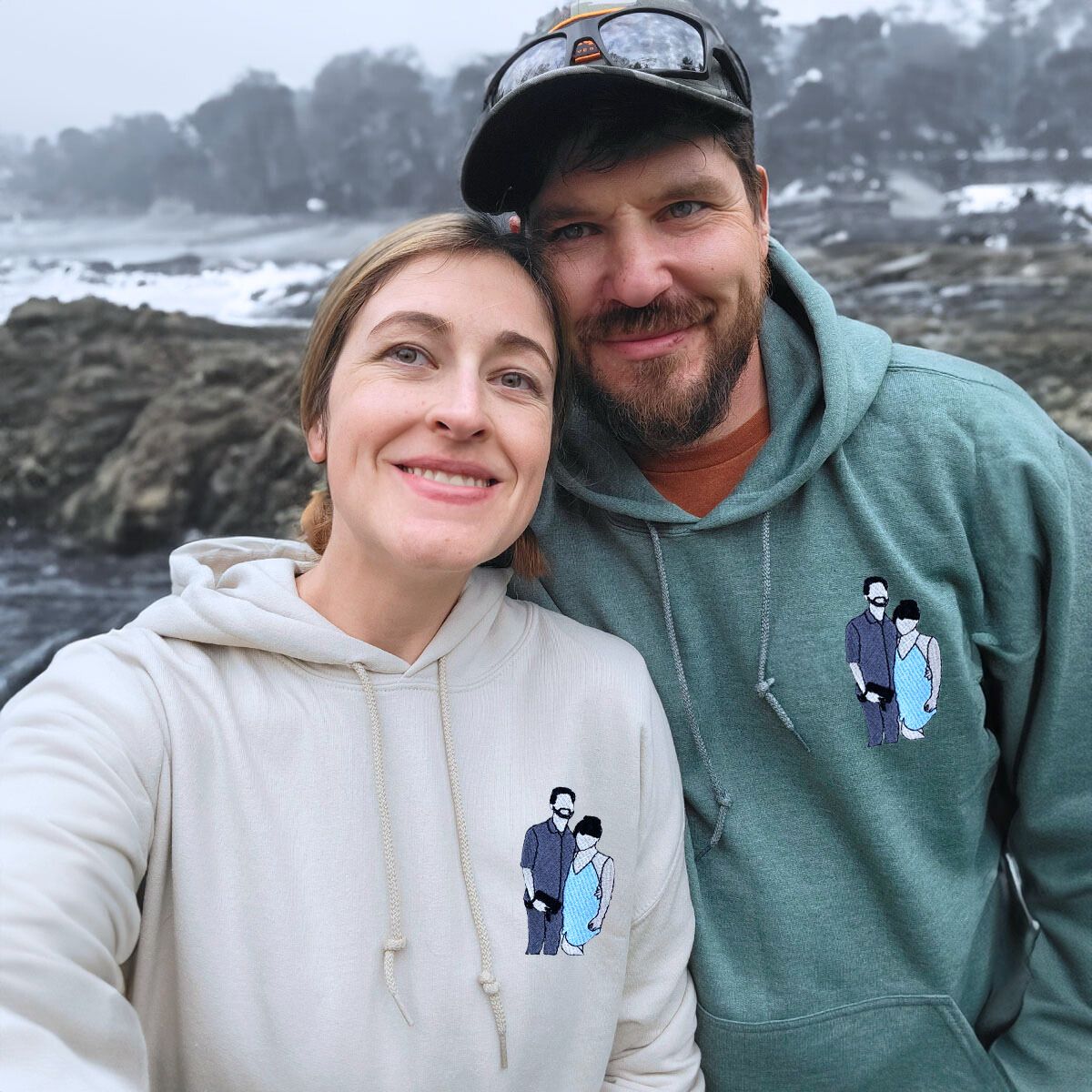 Happy couple near the ocean, wearing personalized beige and green Embroly hoodies with embroidered artwork.