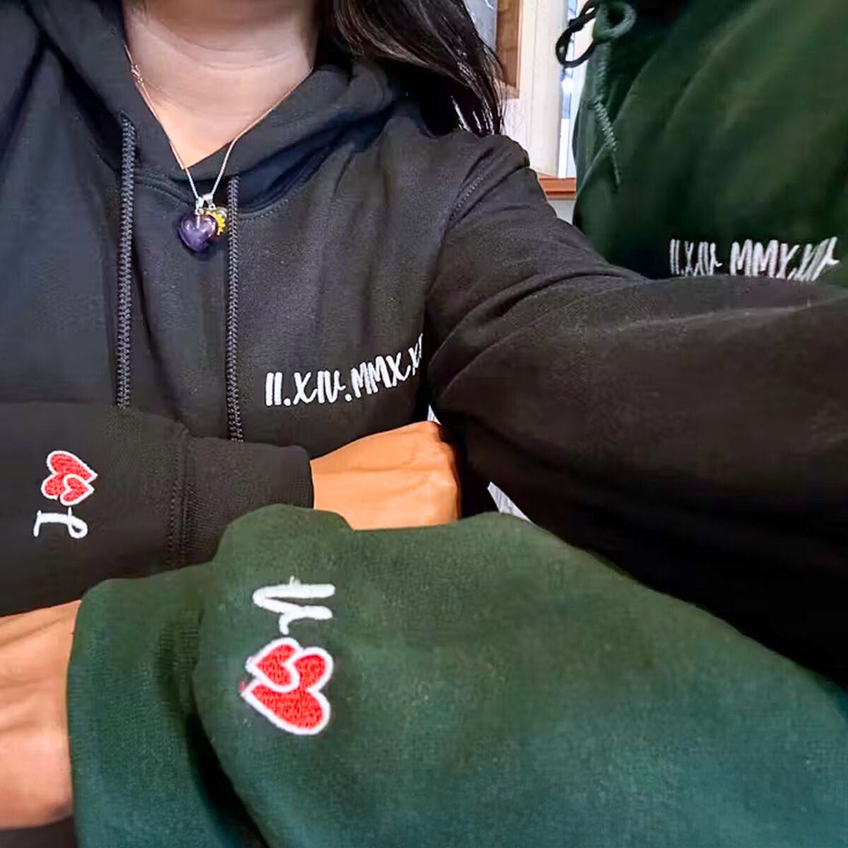 Close-up of a couple wearing matching black and green Embroly hoodies with personalized date and heart embroidery.