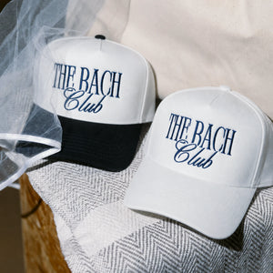 Two white trucker-style baseball caps with navy blue embroidered text reading 'THE BACH Club' are styled on a textured gray herringbone fabric with a beige fabric backdrop and a sheer white veil.