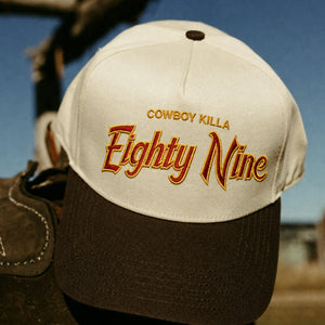 A close-up of a baseball cap with a cream crown and dark brown brim. The front is embroidered with the words 'COWBOY KILLA Eighty Nine' in gold and red lettering.