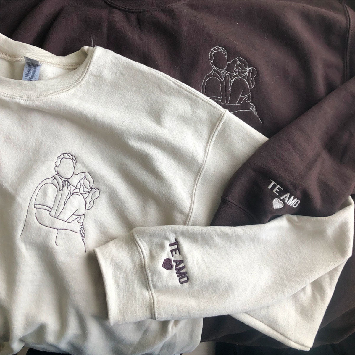 Cream and brown sweatshirts with hand-drawn couple portrait embroidery and "Te Amo" heart detail on the cuff