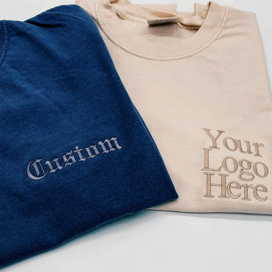 Navy and beige t-shirts folded together, embroidered with gothic-script "Custom" text and a tonal logo placeholder