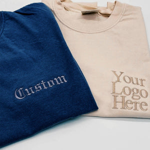 Navy and beige t-shirts folded together, embroidered with gothic-script "Custom" text and a tonal logo placeholder