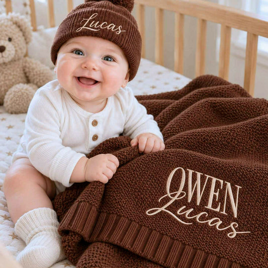 Smiling baby holding a personalized chocolate brown knit blanket with embroidered names and wearing a matching beanie, a gift idea for a baby shower.