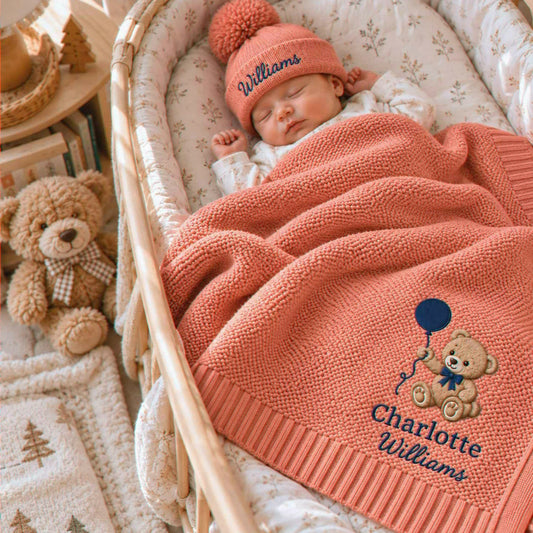Sleeping baby in a coral knit blanket embroidered with a teddy bear and two names, wearing a matching personalized beanie, a gift for a newborn arrival.