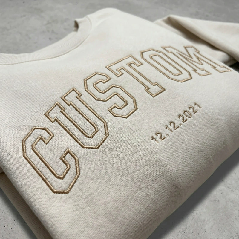 Close-up of tan embroidery thread on a cream sweatshirt showing custom text and date details.