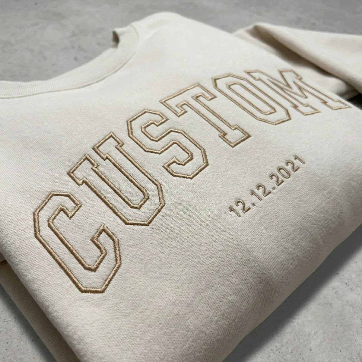 Close-up of tan embroidery thread on a cream sweatshirt showing custom text and date details.