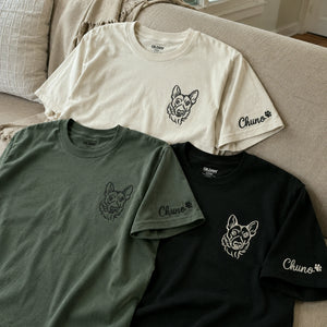 Three short-sleeve crewneck t-shirts laid out on a beige sofa: one cream, one sage green, and one black. Each shirt has a small dog face graphic on the left chest, 'Chuno' text and paw print on the right sleeve, and an OLIMN neck label.