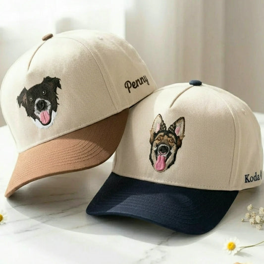 Two personalized baseball caps displayed on a light marble surface with soft neutral background. One cap is beige with a tan brim and an embroidered black-and-white dog face; the other cap is beige with a navy brim and an embroidered brown-and-tan dog face.