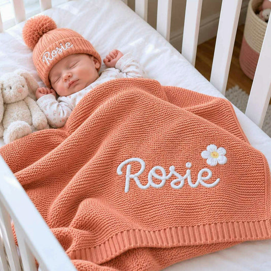 Sleeping newborn wrapped in a peach knit blanket embroidered with a name and white daisy, wearing a matching beanie, a personalized baby shower gift.