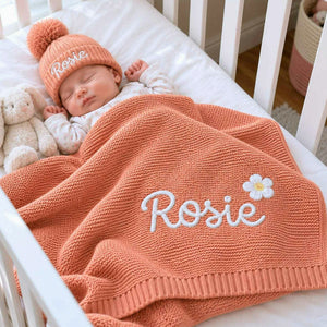 Sleeping newborn wrapped in a peach knit blanket embroidered with a name and white daisy, wearing a matching beanie, a personalized baby shower gift.