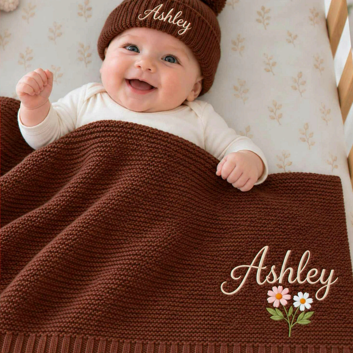 Smiling baby under a personalized chocolate brown knit blanket with embroidered name and flowers, wearing a matching beanie, a gift for a baby shower.