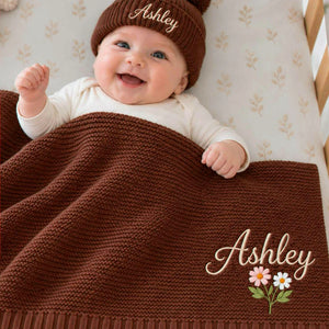 Smiling baby under a personalized chocolate brown knit blanket with embroidered name and flowers, wearing a matching beanie, a gift for a baby shower.