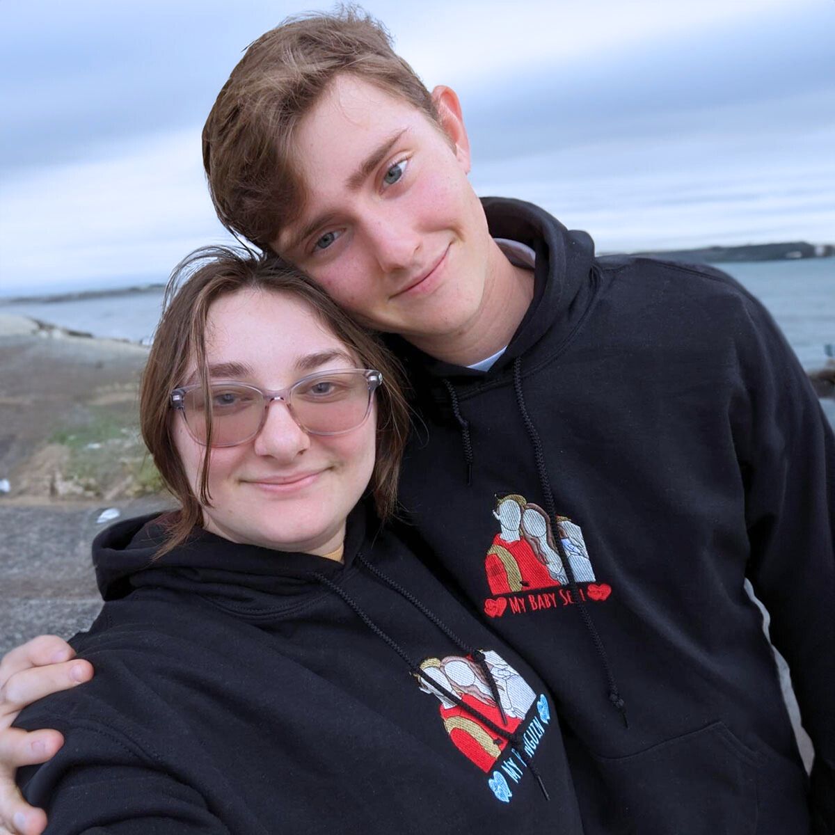 Couple embracing by the seaside, wearing matching black Embroly portrait hoodies with custom embroidery.