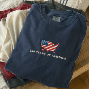 Stack of folded t-shirts with a blue shirt featuring an American flag and text on a wooden surface.
