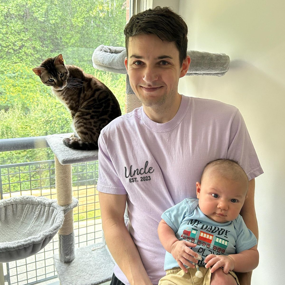 Man in a lavender 'Uncle Est. 2023' embroidered T-shirt holding a baby, celebrating his uncle status.