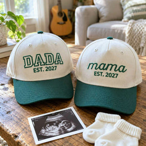 Matching khaki and green Dada and Mama embroidered caps with an established year, a pregnancy announcement gift for expecting parents.