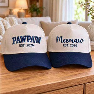 A pair of matching baseball hats on a wooden table. The left hat, in khaki and navy blue, is embroidered with "PAWPAW EST. 2026". The right hat, matching, has "Meemaw EST. 2026" in a script font.