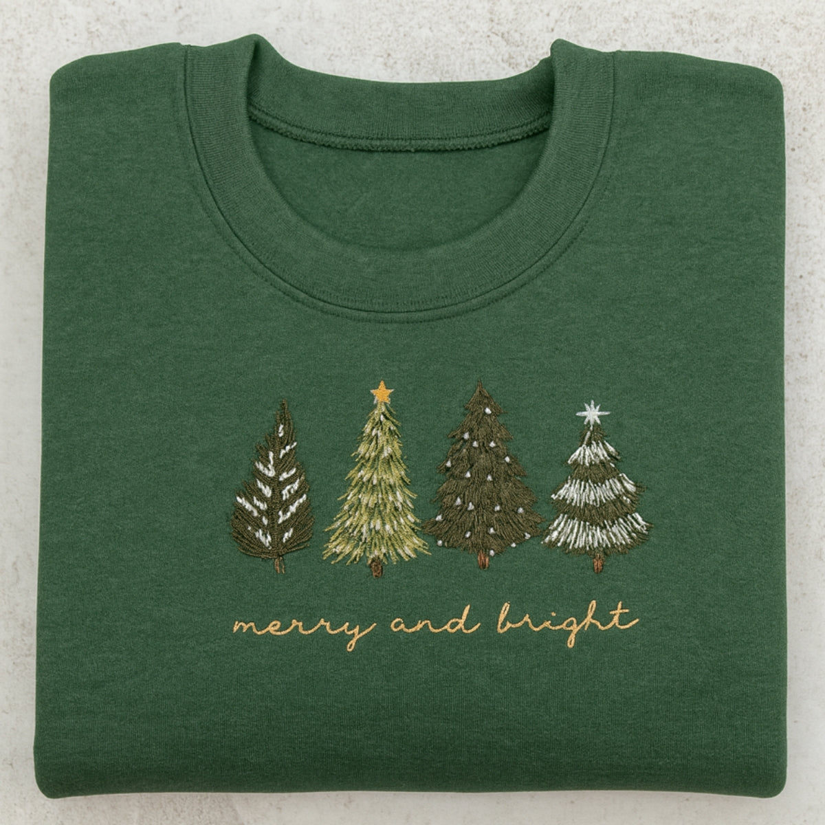 Folded green sweatshirt with embroidered Christmas trees and 'merry and bright' text on a light gray background