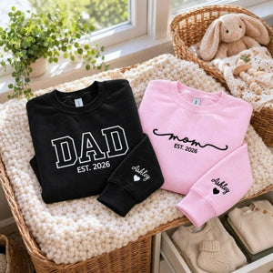 Matching black Dad and light pink Mom embroidered sweatshirts with personalized sleeve names and hearts, a baby shower gift for new parents.