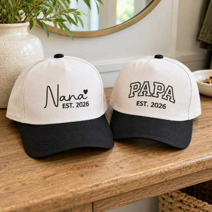 Custom embroidered Nana and Papa Est. 2026 baseball hats set, personalized gifts for new grandparents on a wooden tabletop