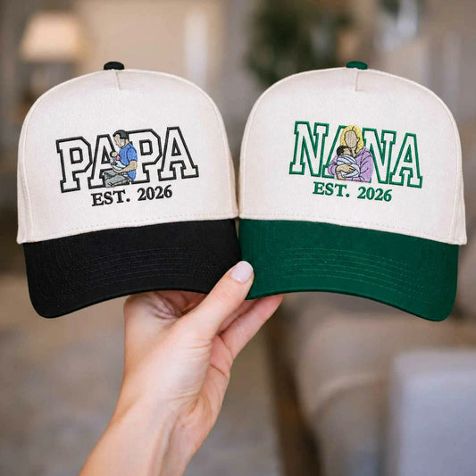 Custom 'Papa EST. 2026' and 'Nana EST. 2026' matching baseball hats on a wooden table, next to a photo of new parents and baby. Personalized grandparent announcement gifts.