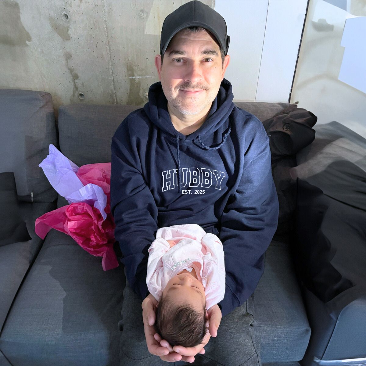 New dad wearing a navy 'Hubby EST. 2025' hoodie, lovingly holding his newborn baby. A thoughtful gift for first-time fathers.