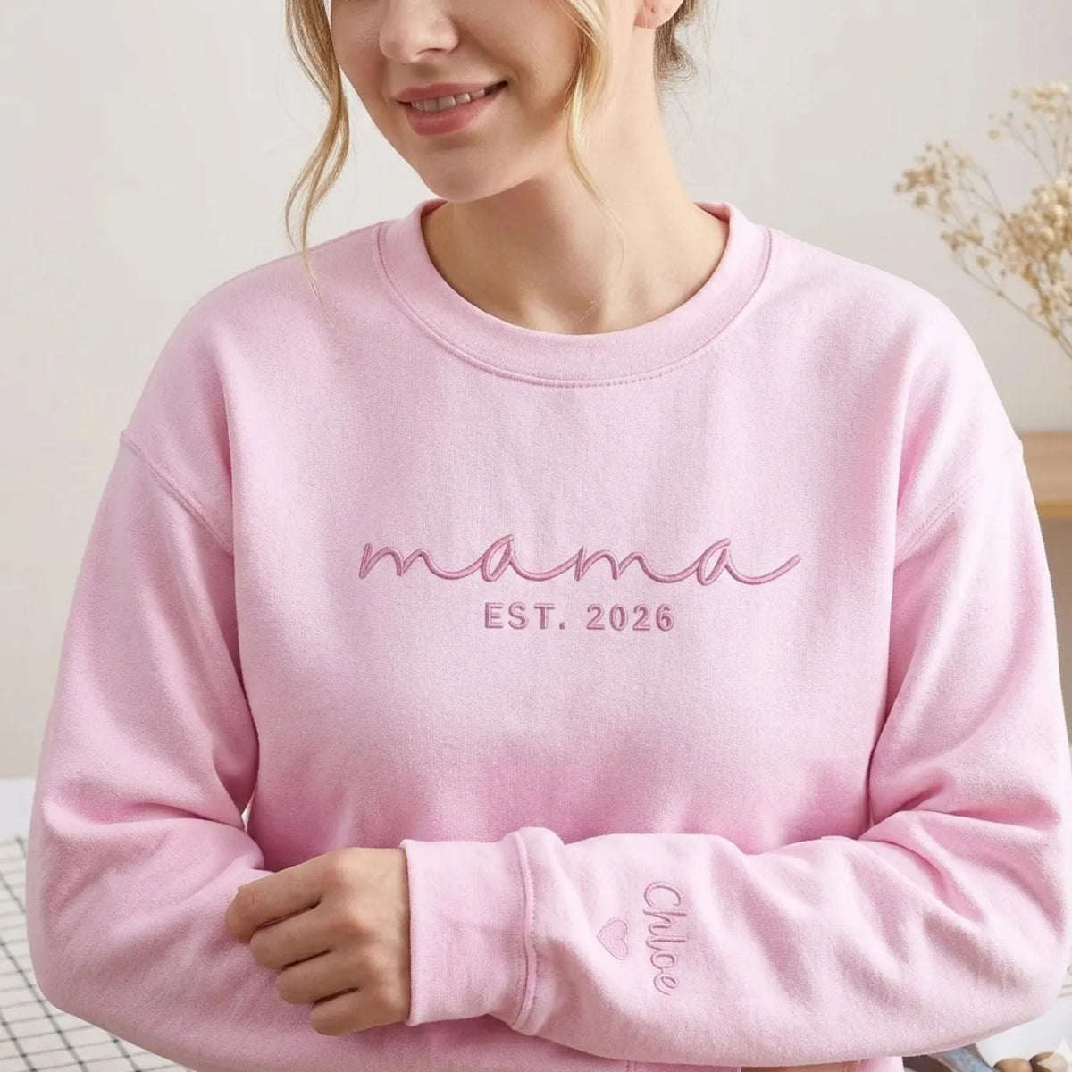 Happy new mom wearing a pink sweatshirt with 'mama EST. 2026' text.