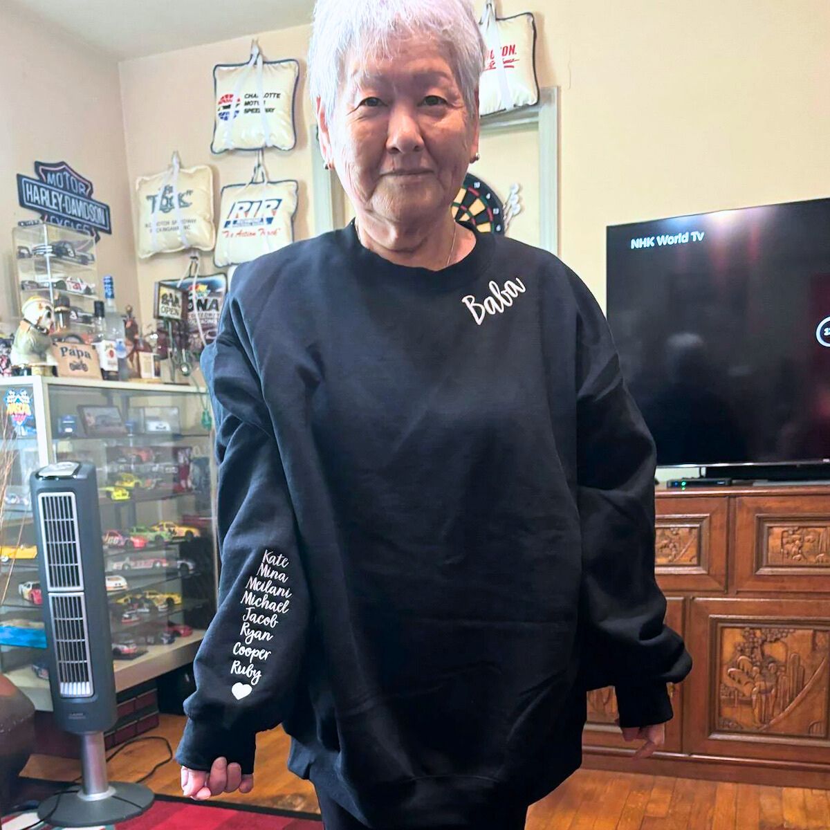 Elderly woman in a black 'Baba' sweatshirt with embroidered names on the sleeve, standing in a room filled with collectibles.