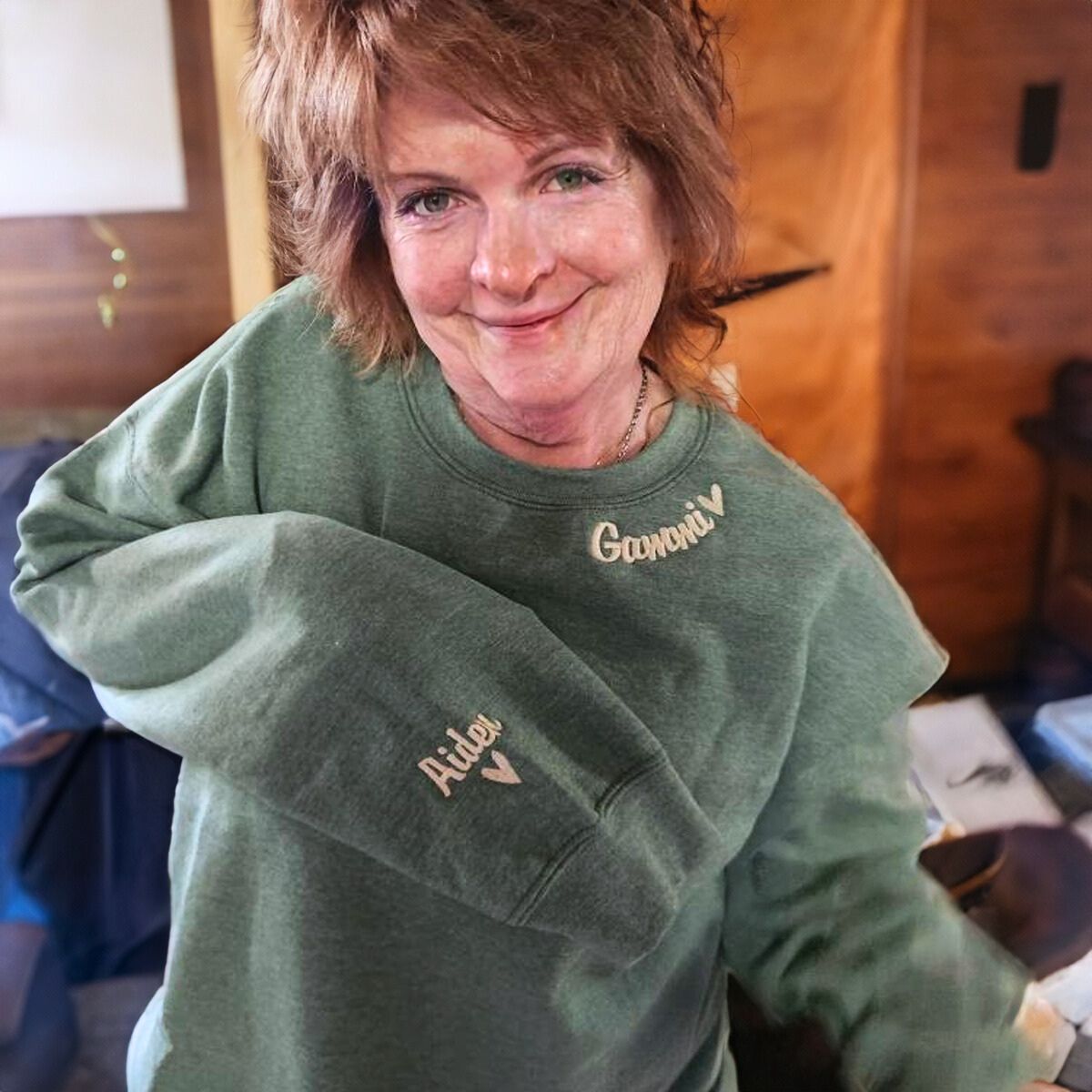 Smiling grandmother in a sage green 'Gamoni' sweatshirt with a personalized embroidered name, posing in a cozy indoor setting.