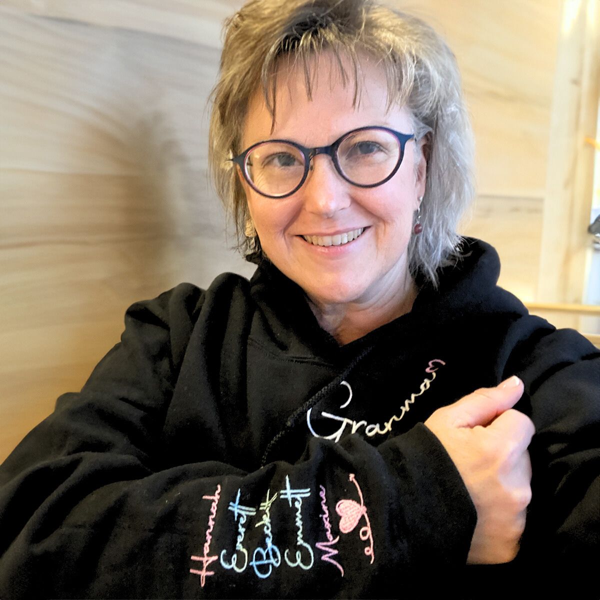 Grandmother in a black 'Grandma' hoodie with cursive embroidery and colorful grandkids' names on the sleeve, posing indoors.