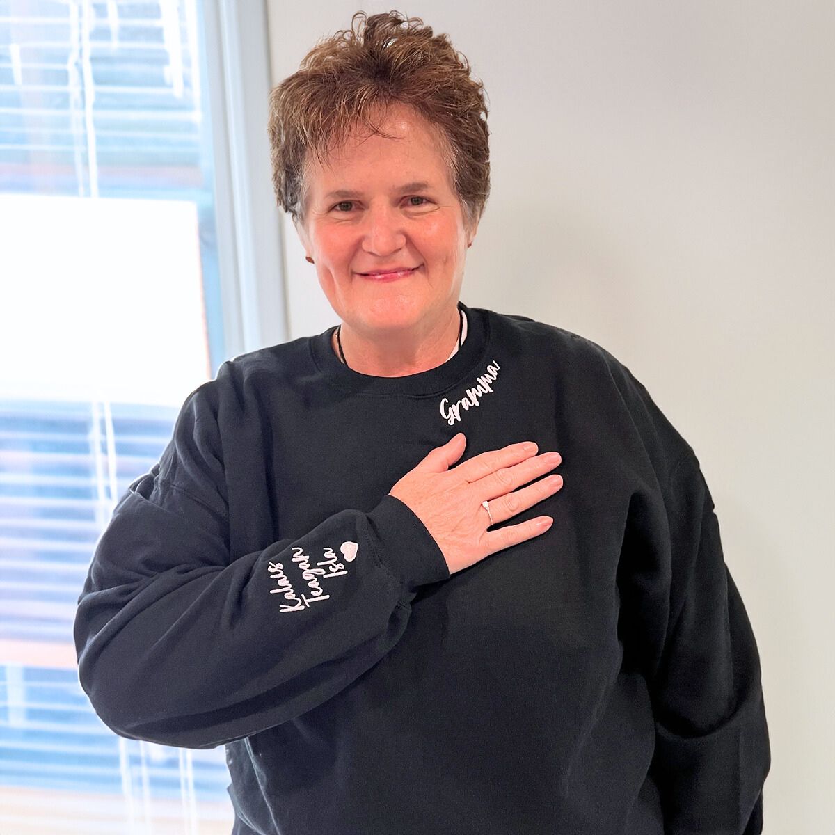 Grandmother in a black 'Grandma' sweatshirt with white embroidery, placing her hand over her heart in a bright indoor setting