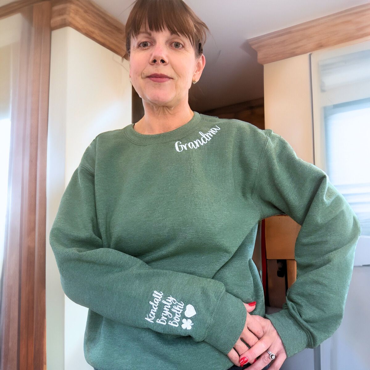 Grandmother with short brown hair in a sage green 'Grandma' sweatshirt, showing white embroidered text and grandkids' names on the sleeve.