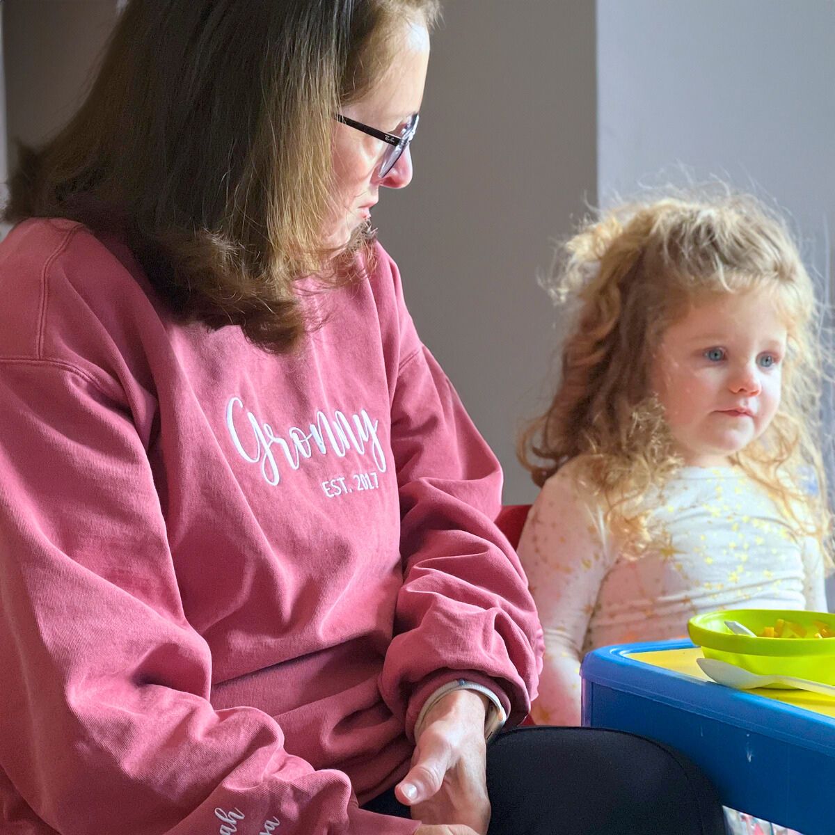 Grandmother wearing a vintage red 'Granny EST. 2017' sweatshirt with white embroidered text, sitting with her curly-haired granddaughter at a small table.