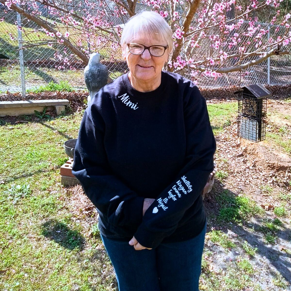 Elderly woman in a black 'Mimi' sweatshirt with embroidered names on the sleeve, standing in a garden with pink blossoms.