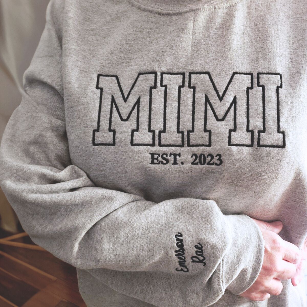 Close-up of a light gray 'MIMI EST. 2023' sweatshirt with bold black embroidered text and a personalized name on the sleeve, held in a cozy indoor setting.